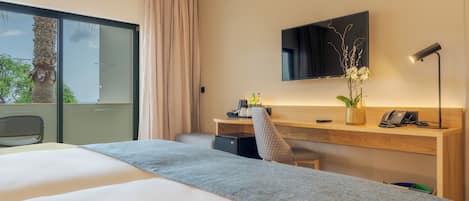 Habitación de hotel con una cama grande, un escritorio con computadora, una silla, televisor montado en la pared y ventana con vista a árboles.