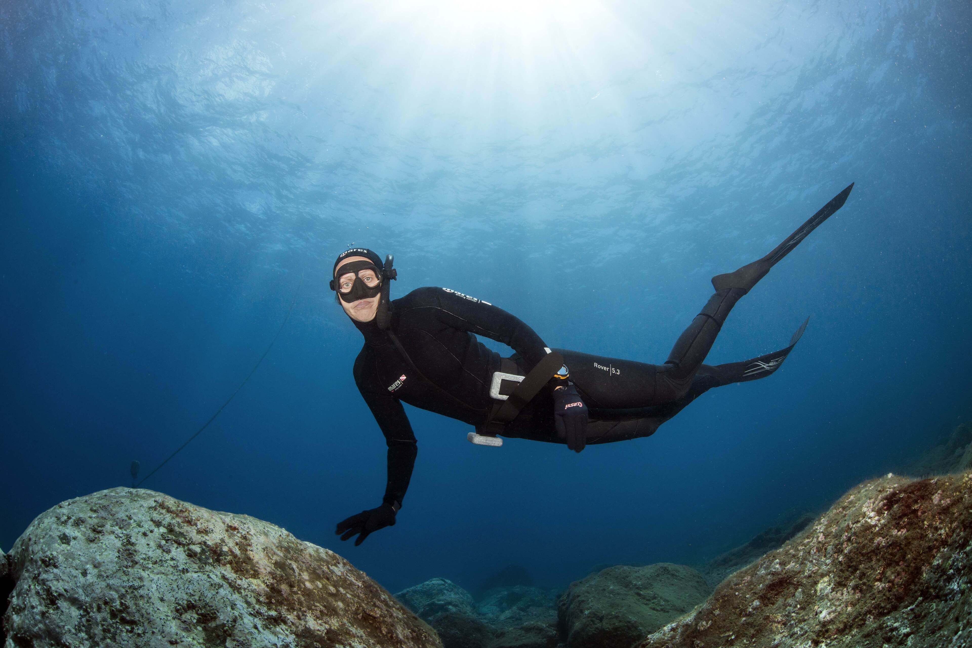 A diver in a black wetsuit with a dive computer and fins, swimming underwater with a rocky seabed and clear blue water.