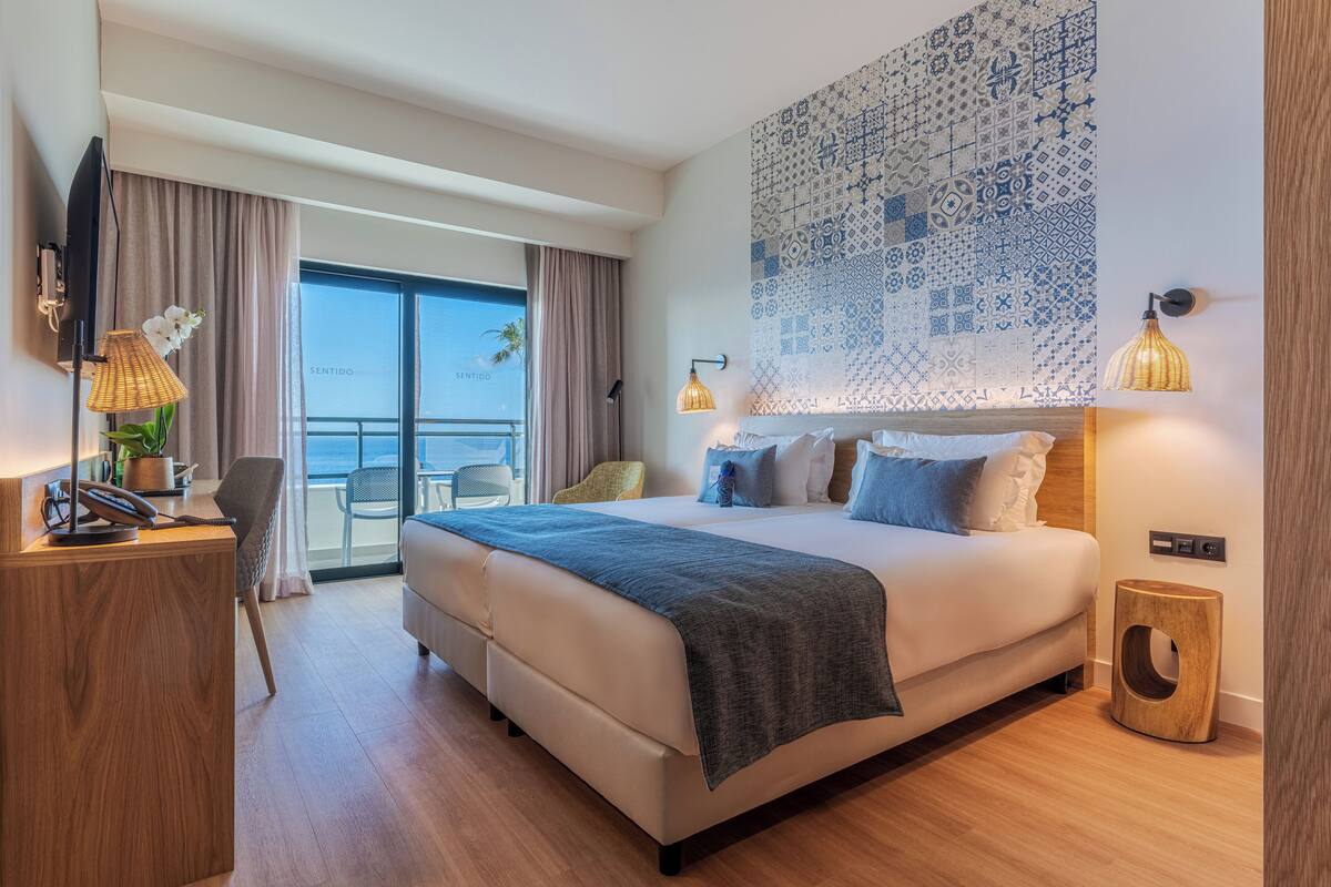 A modern hotel room with a large bed, a desk, a chair, and a view of the sea.
