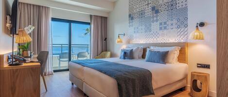A modern hotel room with a large bed, a desk, a chair, and a view of the sea.