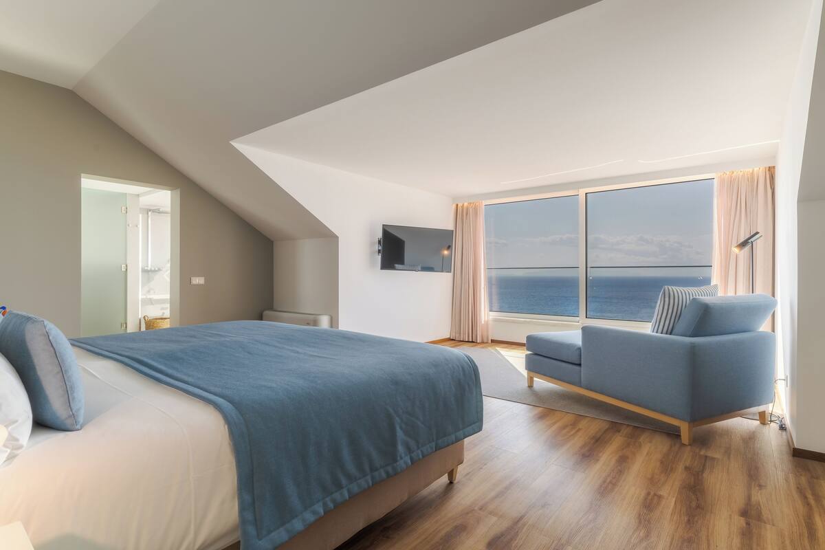 A modern bedroom with a large bed, a blue armchair, and a large window offering a view of the sea.