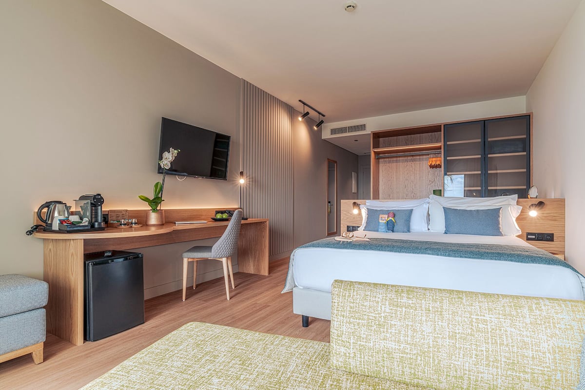 A modern hotel room with a bed, desk, chair, and a small kitchenette.