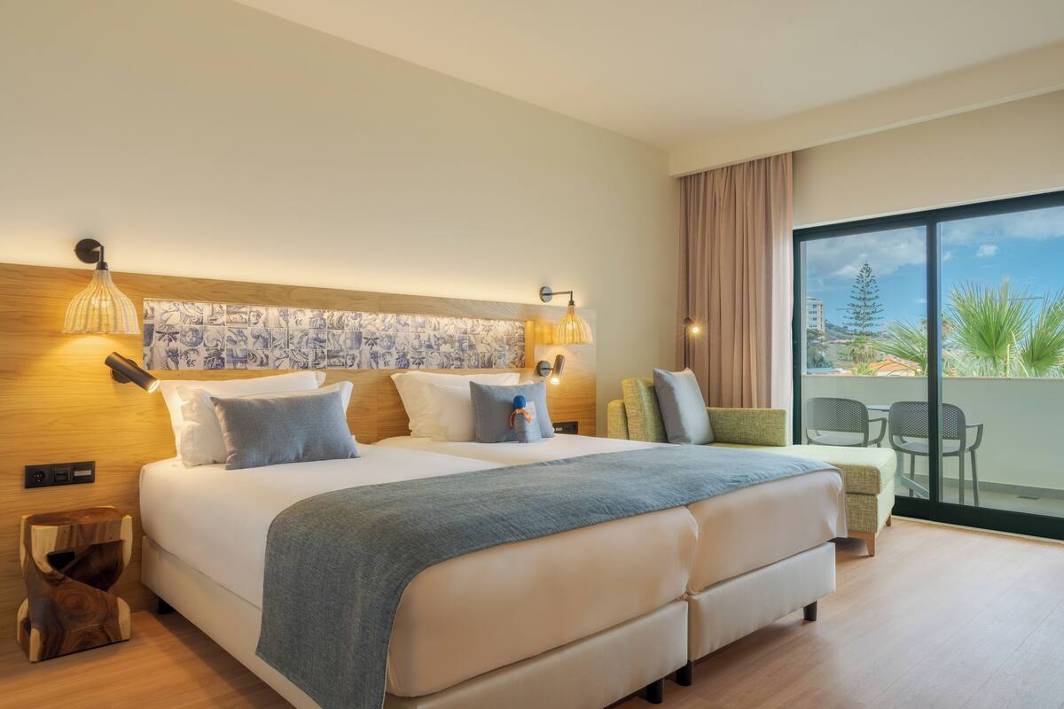A modern hotel room with a large bed, two bedside tables, a nightstand, and a balcony with a view of palm trees and buildings.