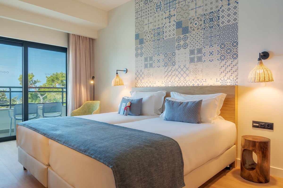 A modern hotel room with a large bed, a balcony with chairs, and a patterned wall.
