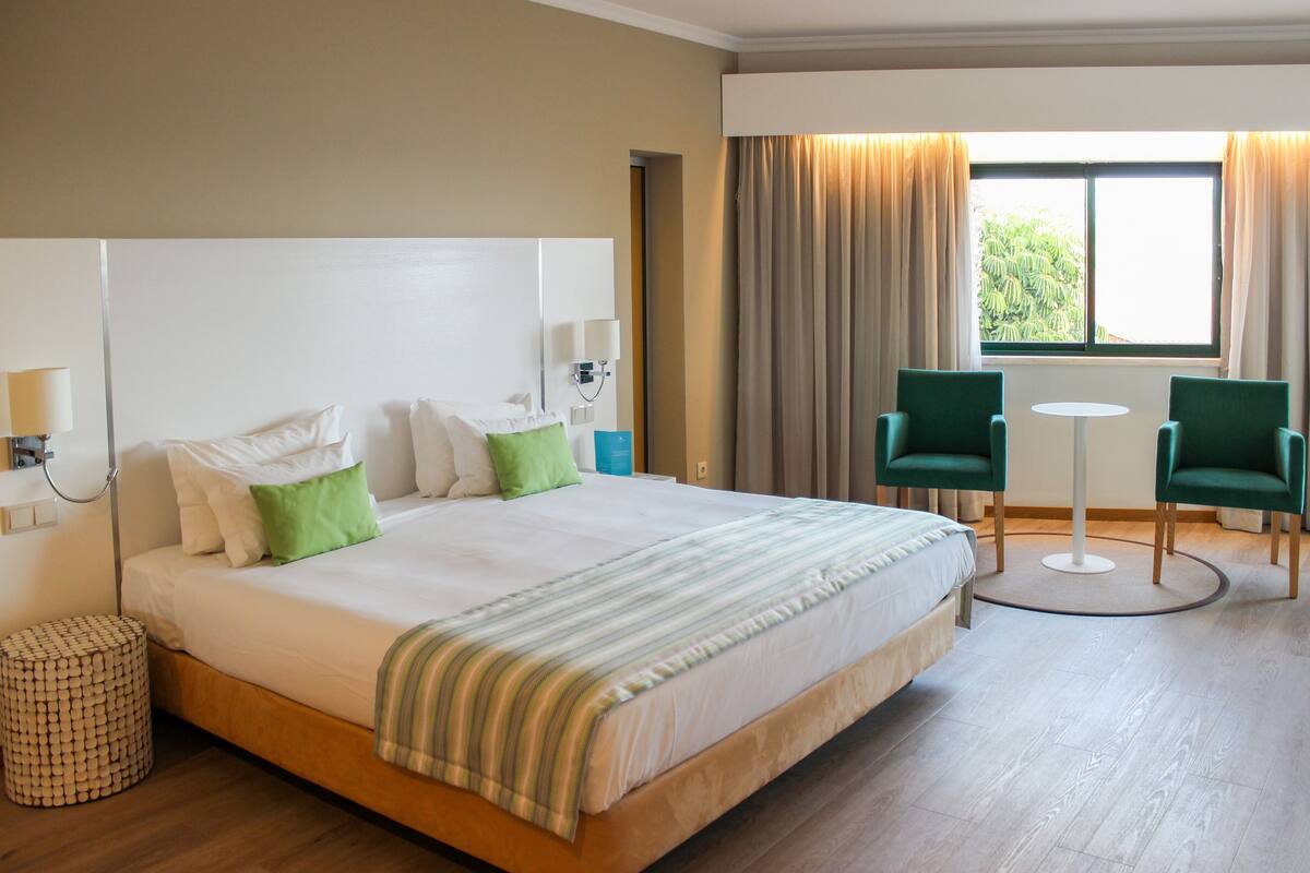 A hotel room with a large bed, two chairs, a small table, and a window with curtains.