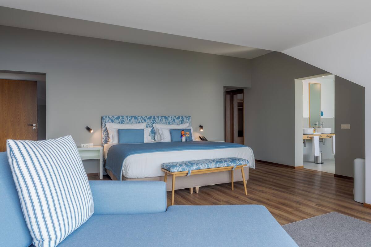 A modern bedroom with a large bed, a bedside table, a bench, a sofa, and a bathroom visible through an open door.