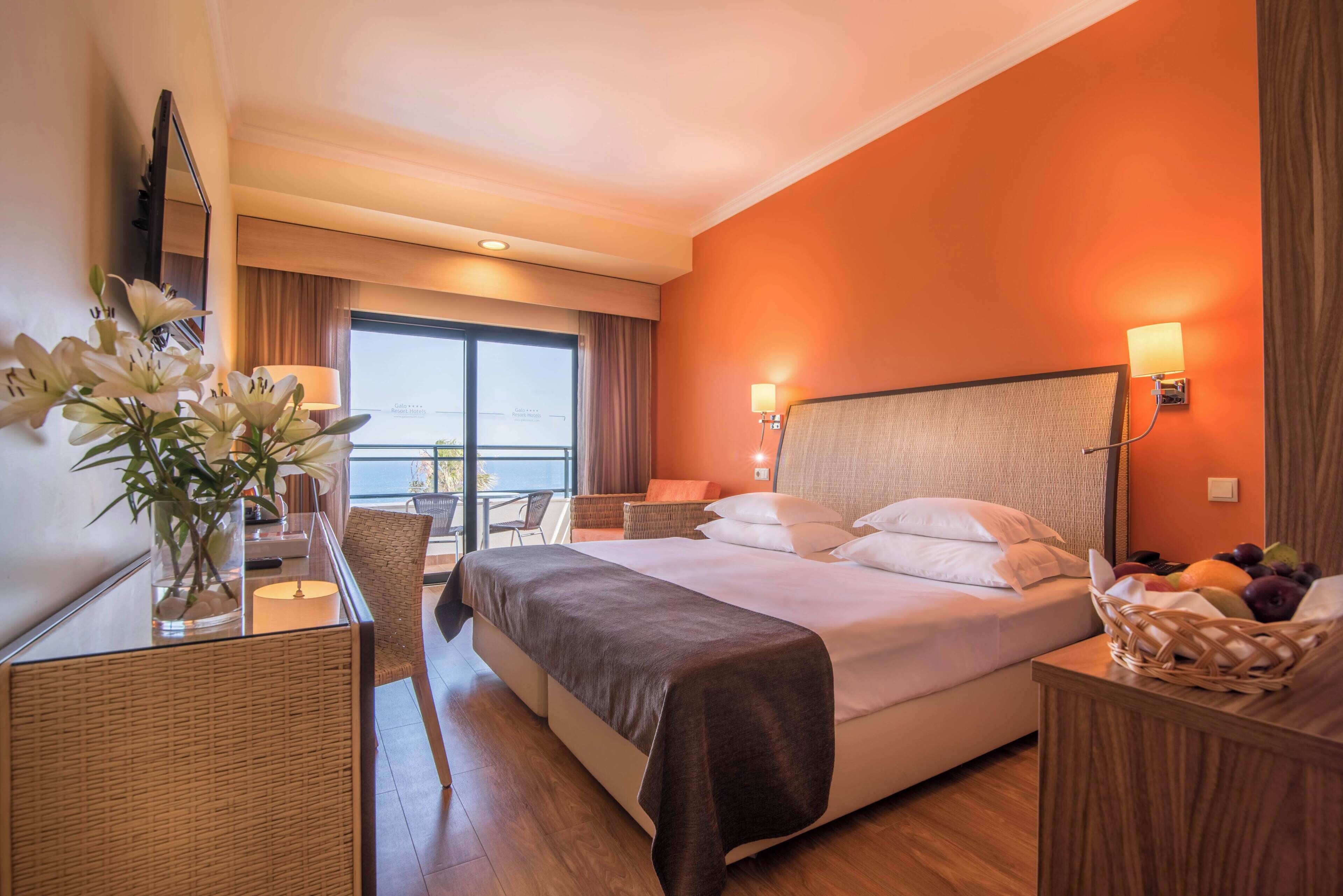 A hotel room with a bed, a desk with a lamp, a chair, a window with curtains, and a balcony.