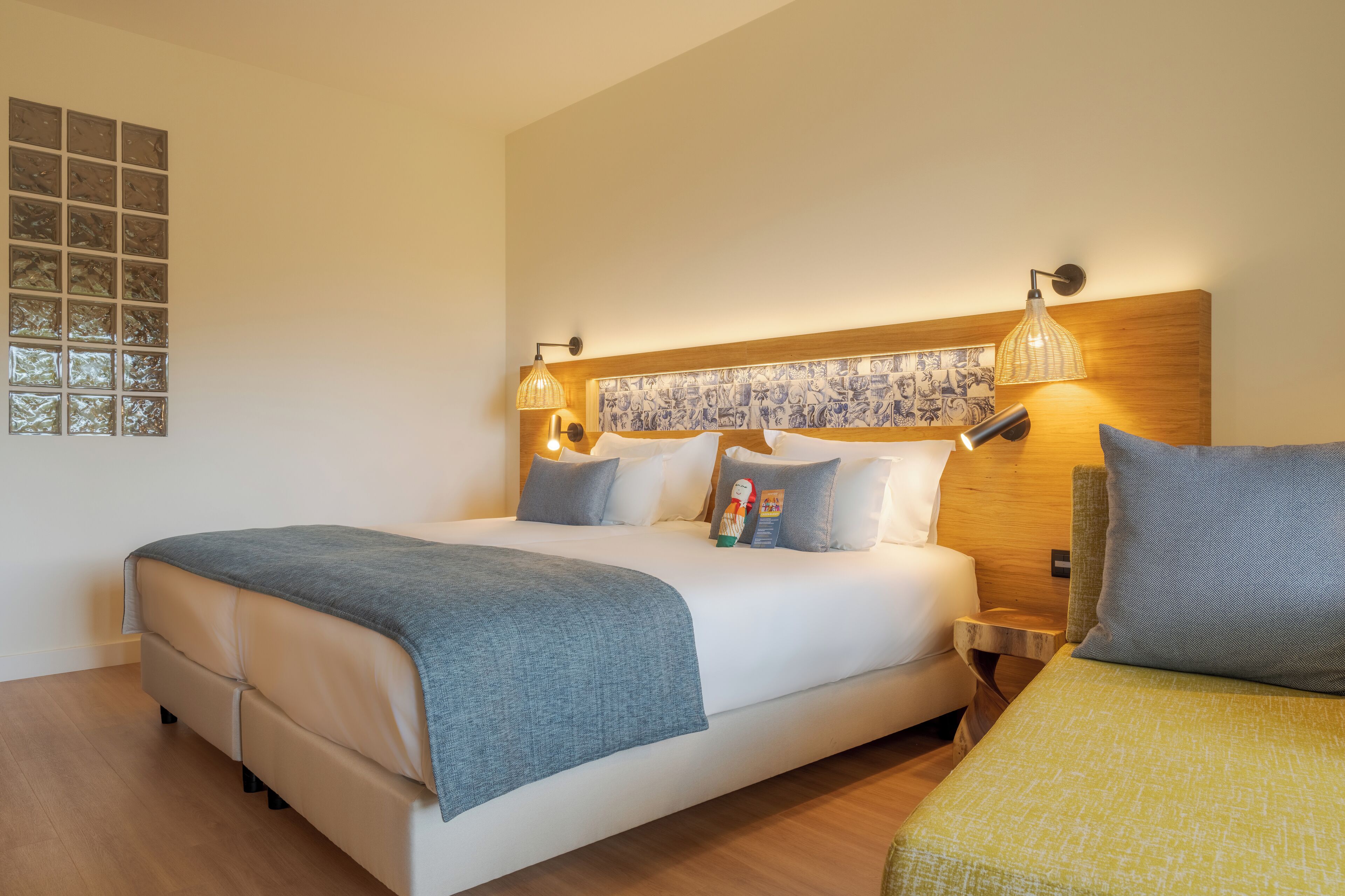 Un lit bien fait, recouvert d’une couverture grise, avec une tête de lit en bois ornée d’un motif décoratif, deux lampes de chevet et une petite table sur laquelle se trouve une lampe.