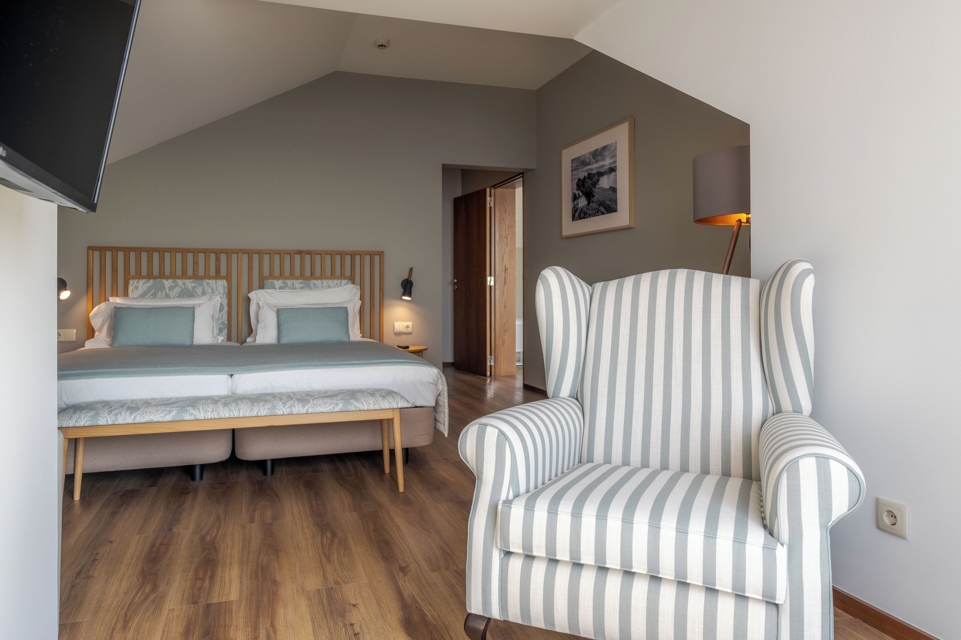 A bedroom with a bed, bedside tables, a chair, a TV, and a door leading to another room.