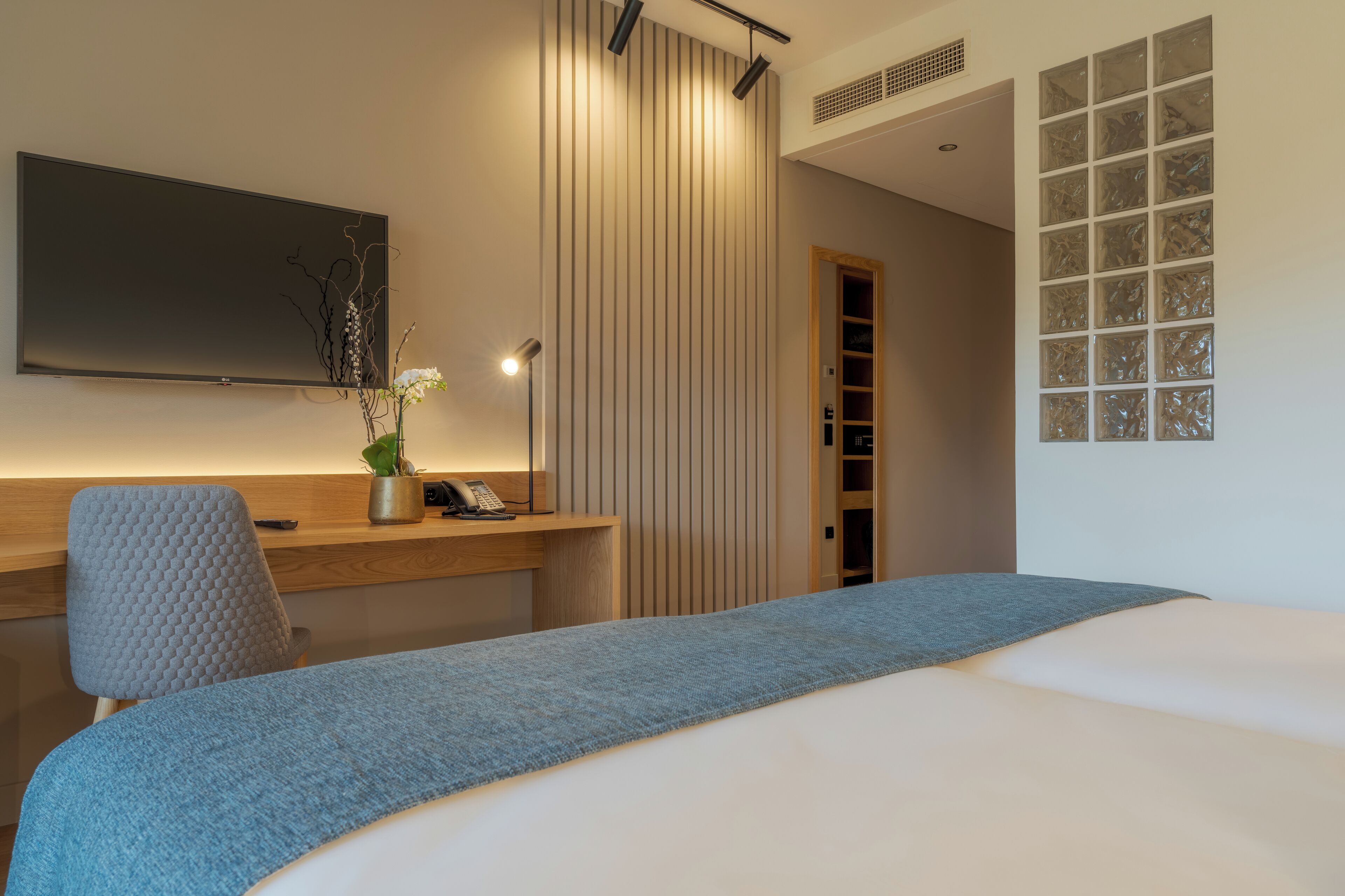 Habitació d'hotel moderna amb un llit, un escriptori amb un ordinador, una cadira, un televisor muntat a la paret i una finestra amb panells de vidre mat.