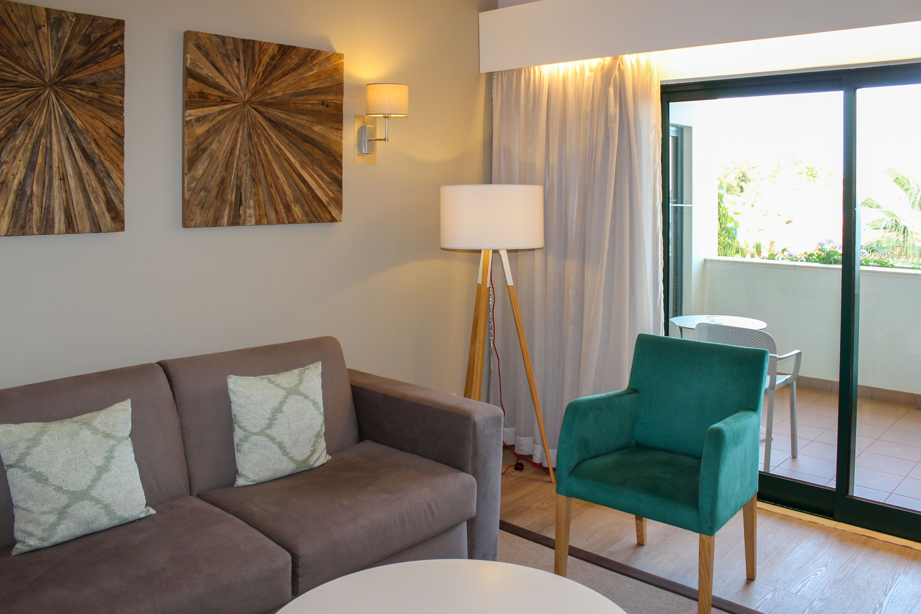 A modern living room with a brown sofa, teal armchair, and a white table. There are two wall-mounted lamps and a large window with curtains.