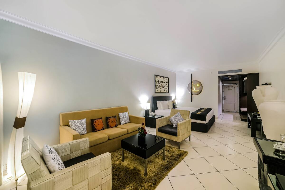 A modern living room with a beige sofa, a black coffee table, and a patterned rug. There is a white wall with a decorative piece and a door leading to another room.