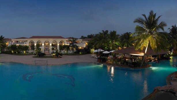 A large outdoor swimming pool surrounded by palm trees and lounge chairs, with a building in the background. - The Zuri White Sands, Goa Resort & Casino (Varca)
