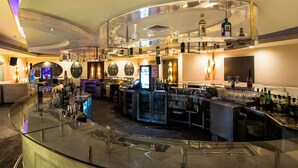 A modern bar area with a curved glass counter, multiple shelves with bottles, and a digital screen displaying content. - The Zuri White Sands, Goa Resort & Casino (Varca)