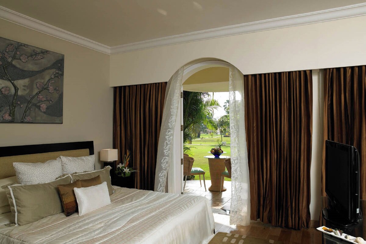 A hotel room with a bed, a painting, a TV, and a view of a garden through an open doorway.