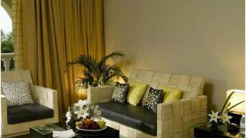 A living room with a beige sofa, two armchairs, a coffee table with a fruit bowl, and a potted plant.