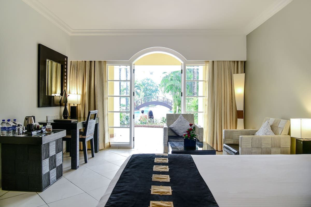 A hotel room with a bed, a desk, a chair, a sofa, and a view of a pool and greenery outside.