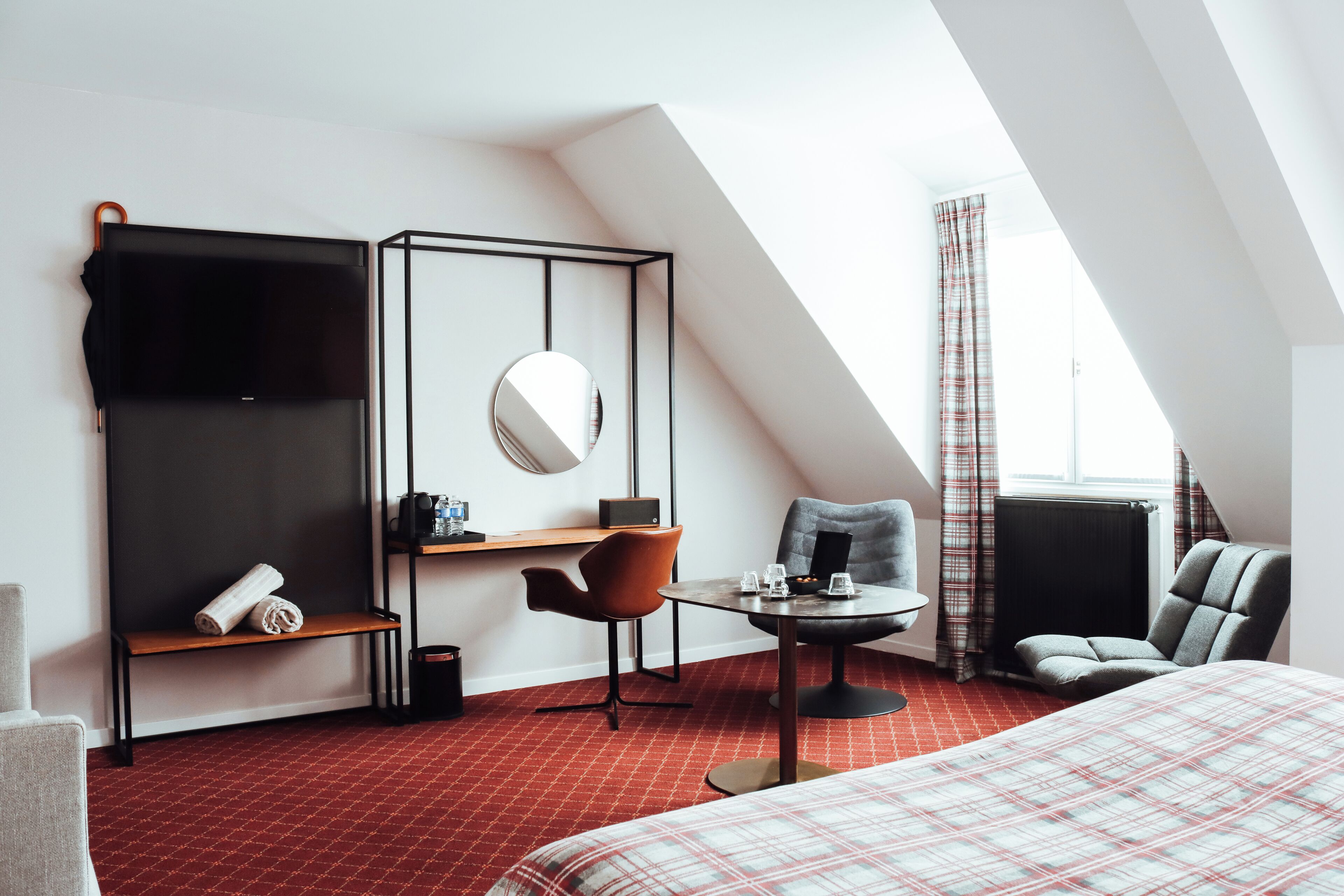 A modern hotel room with a red patterned carpet, a bed, a desk with a chair, a sofa, a TV, and a window with curtains.