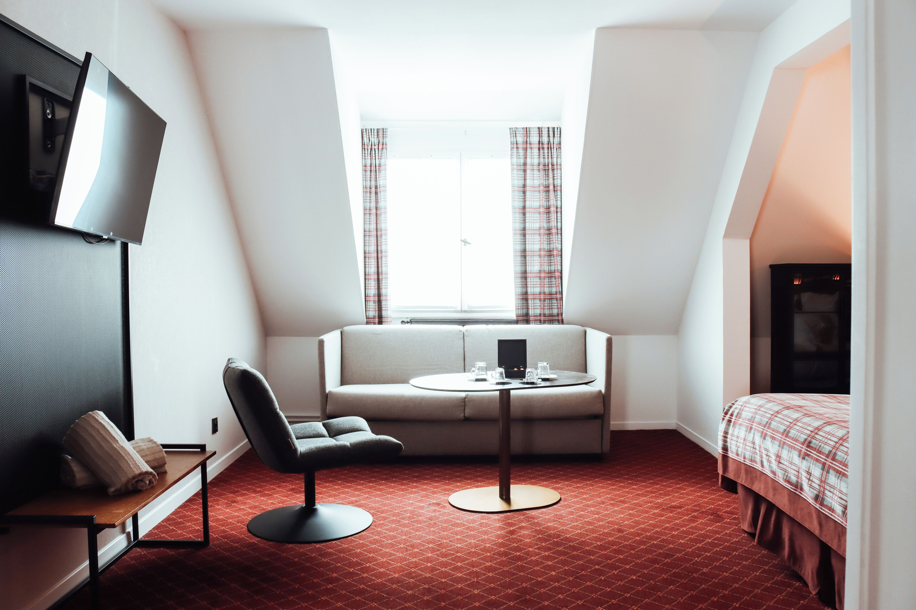 Une chambre d’hôtel compacte comprenant un canapé, une petite table ronde, un téléviseur fixé au mur et un lit avec une couvre-lit à carreaux.