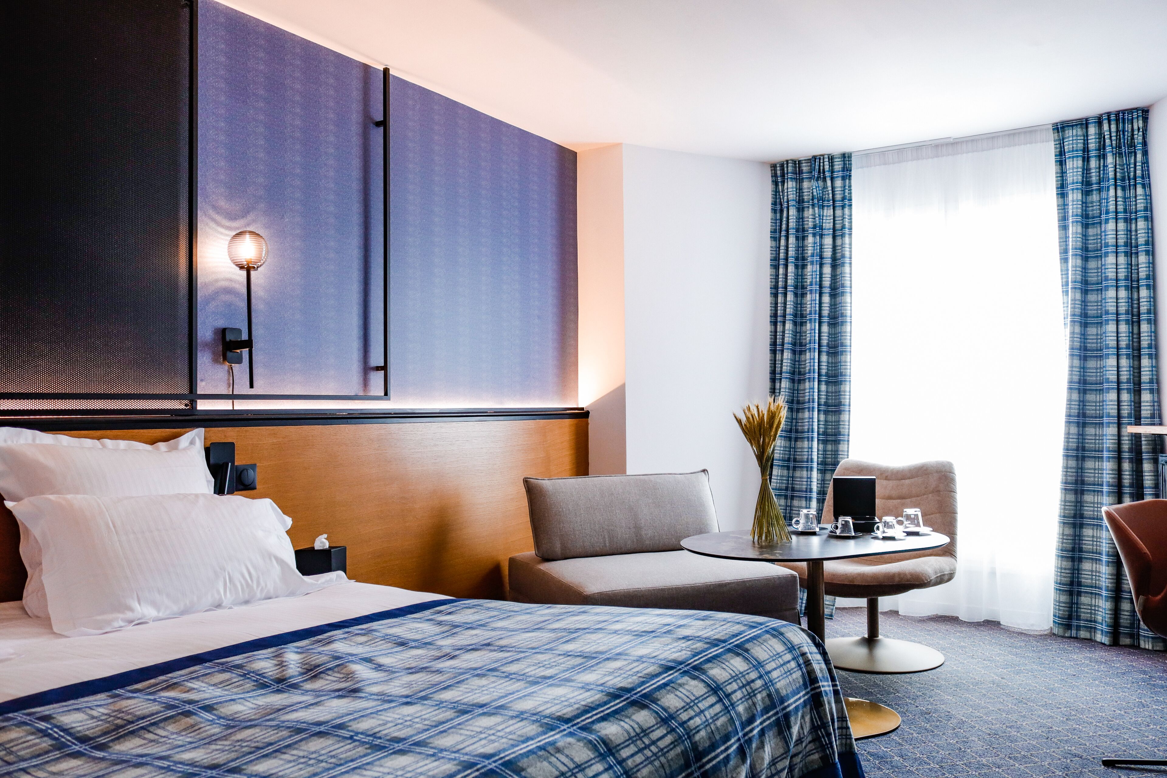 A hotel room with a large bed, a desk with a chair, a small table with a vase, and a window with curtains.