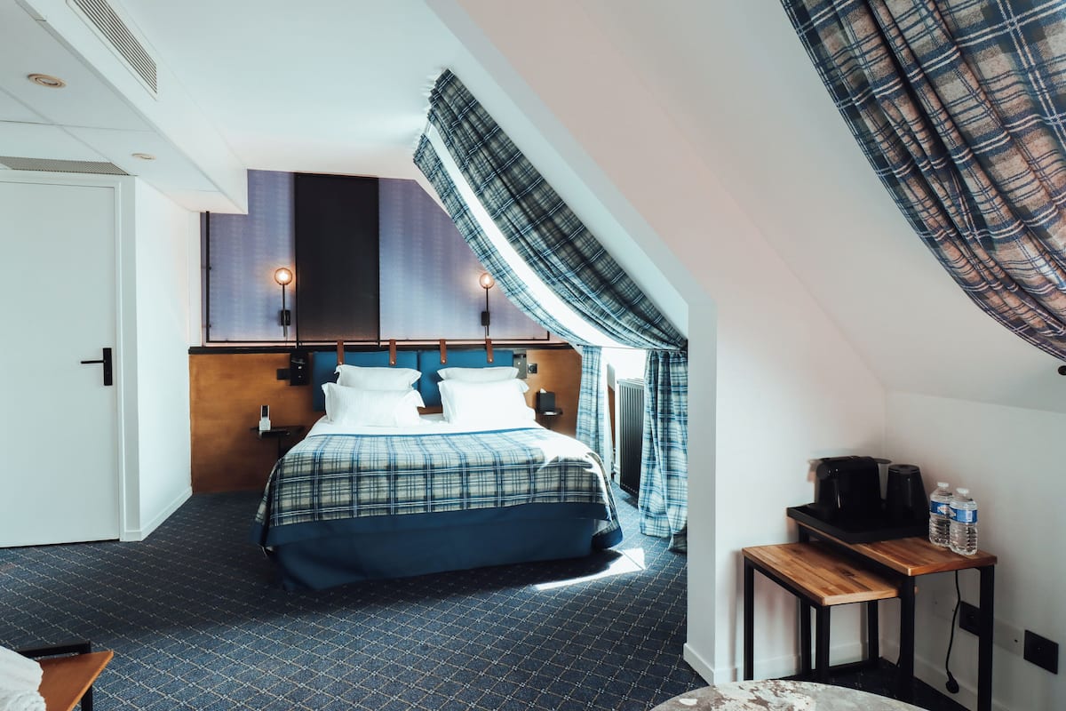 Ein Hotelzimmer mit einem Bett, einem kleinen Tisch mit Wasserkocher und Tassen, einem Heizkörper und einem Fenster mit Vorhängen.