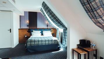 A hotel room with a bed, a small table with a kettle and cups, a radiator, and a window with curtains.