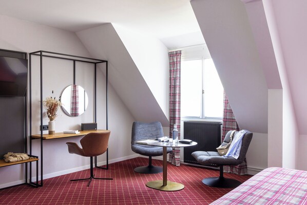 A modern hotel room with a sloped ceiling, a desk with a chair, a round mirror, a small table with a chair, a bed with a plaid bedspread, and a radiator.