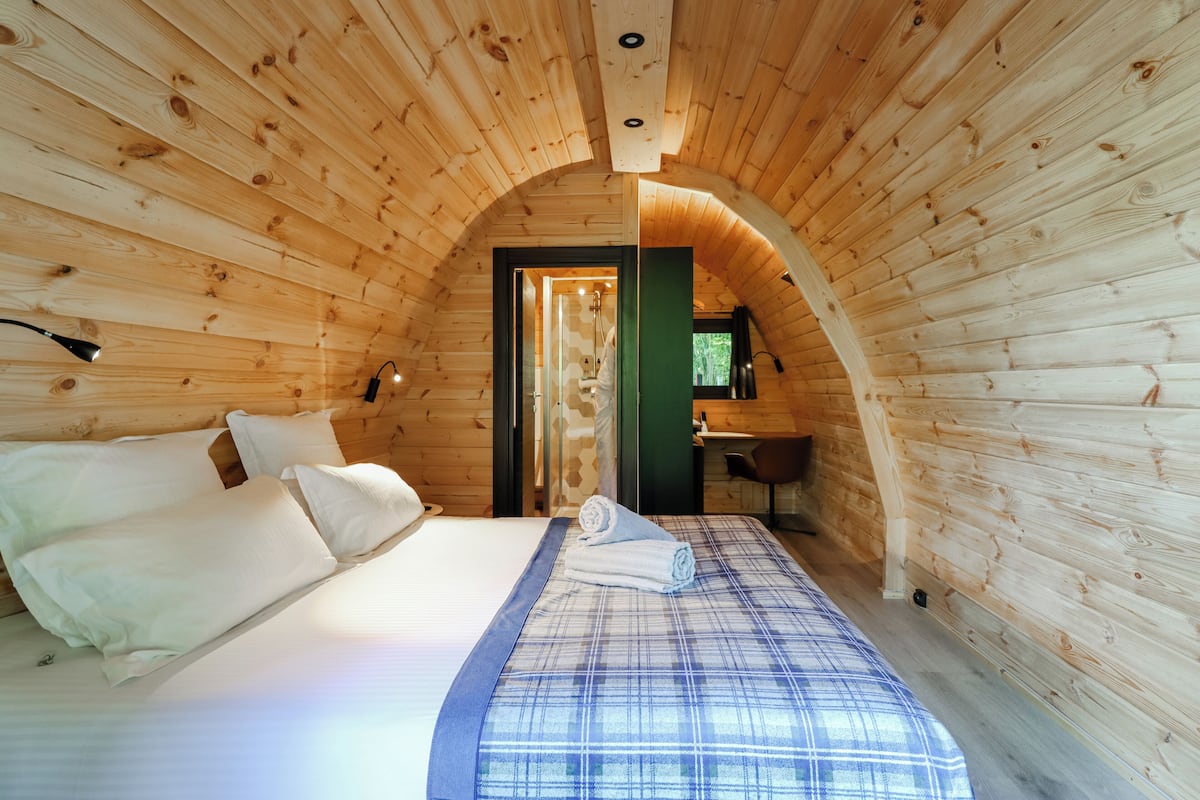 Ein gemütliches Schlafzimmer in einer Holzhütte mit Bett, Kissen, einem aufgerollten Handtuch und einem kleinen Schreibtisch mit Stuhl.