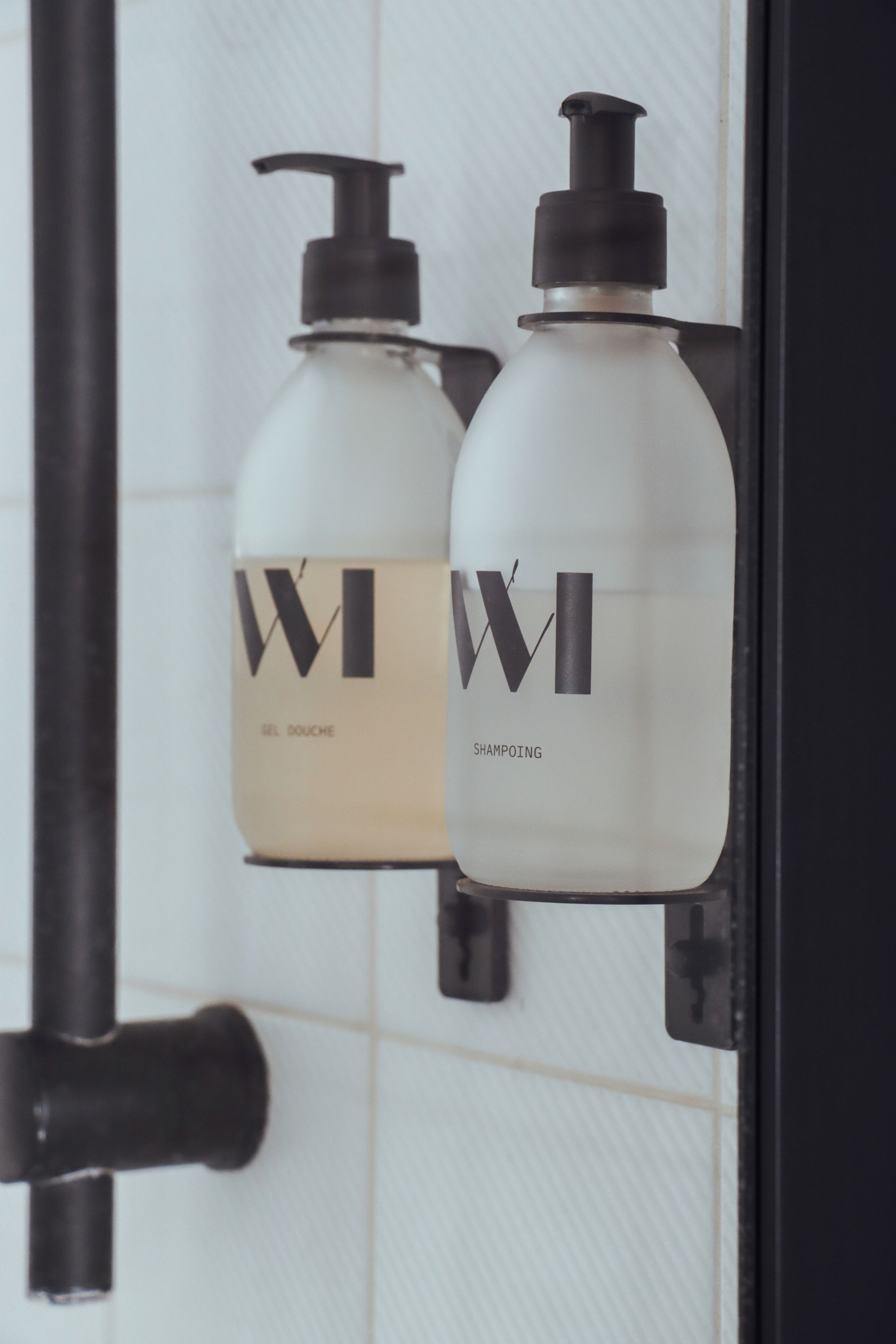 Two white bottles with black pumps labeled 'WI' and 'WI SHAMPOOING' mounted on a tiled wall.