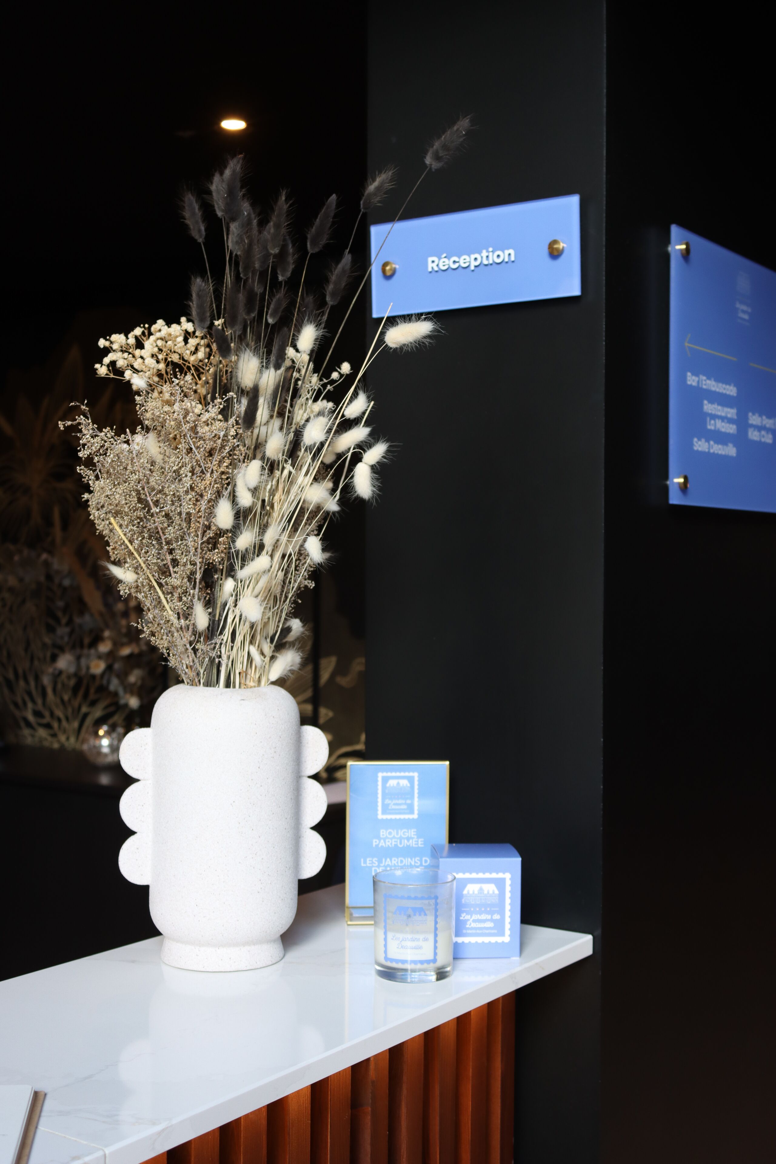 Een receptiebalie met een witte vaas met gedroogde bloemen, een blauw bordje dat het receptiegebied aangeeft, en een klein presentatiemodel van parfumflesjes.