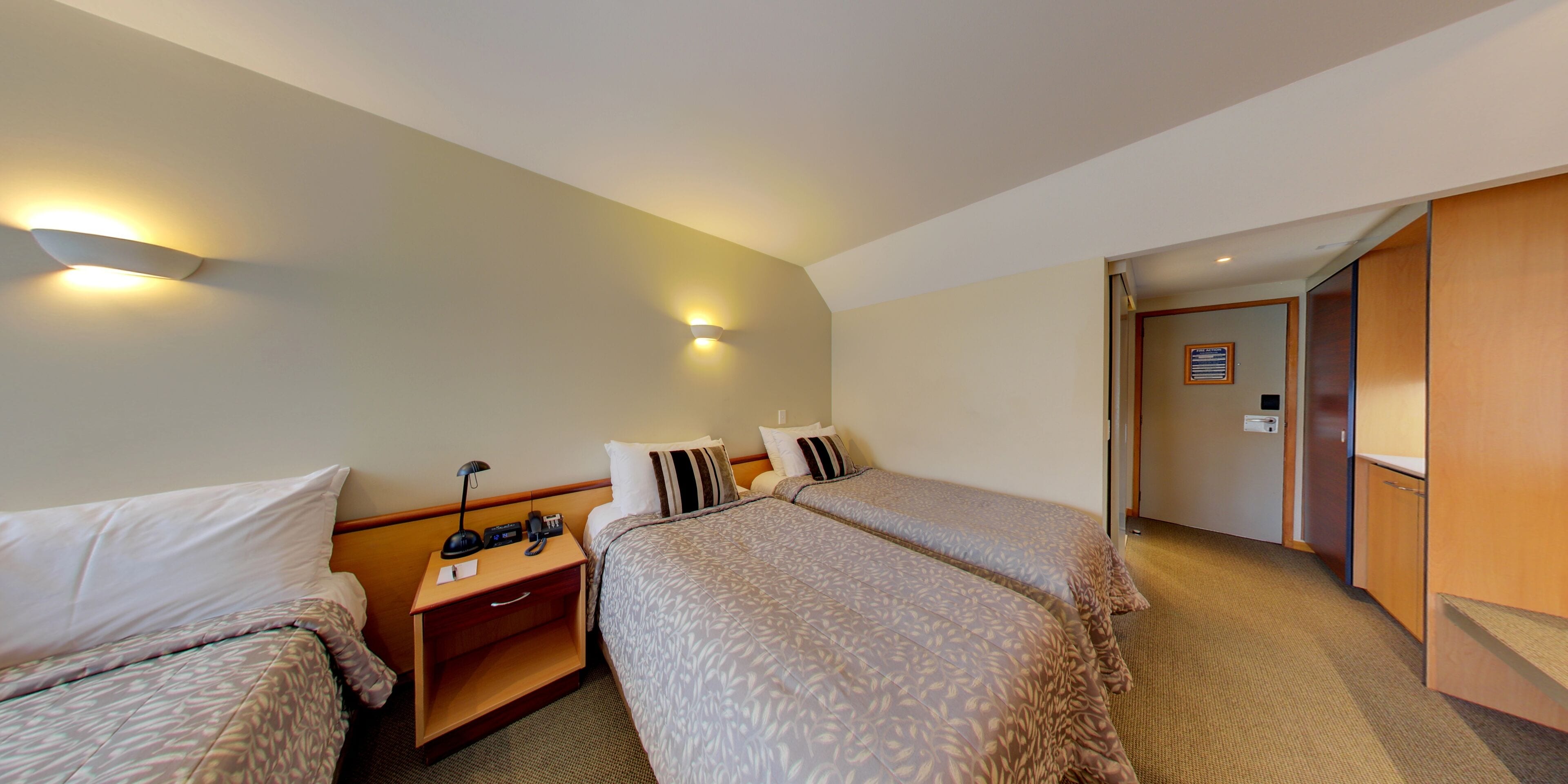 A hotel room with two beds, a nightstand with a lamp, a telephone, and a wall-mounted light fixture.