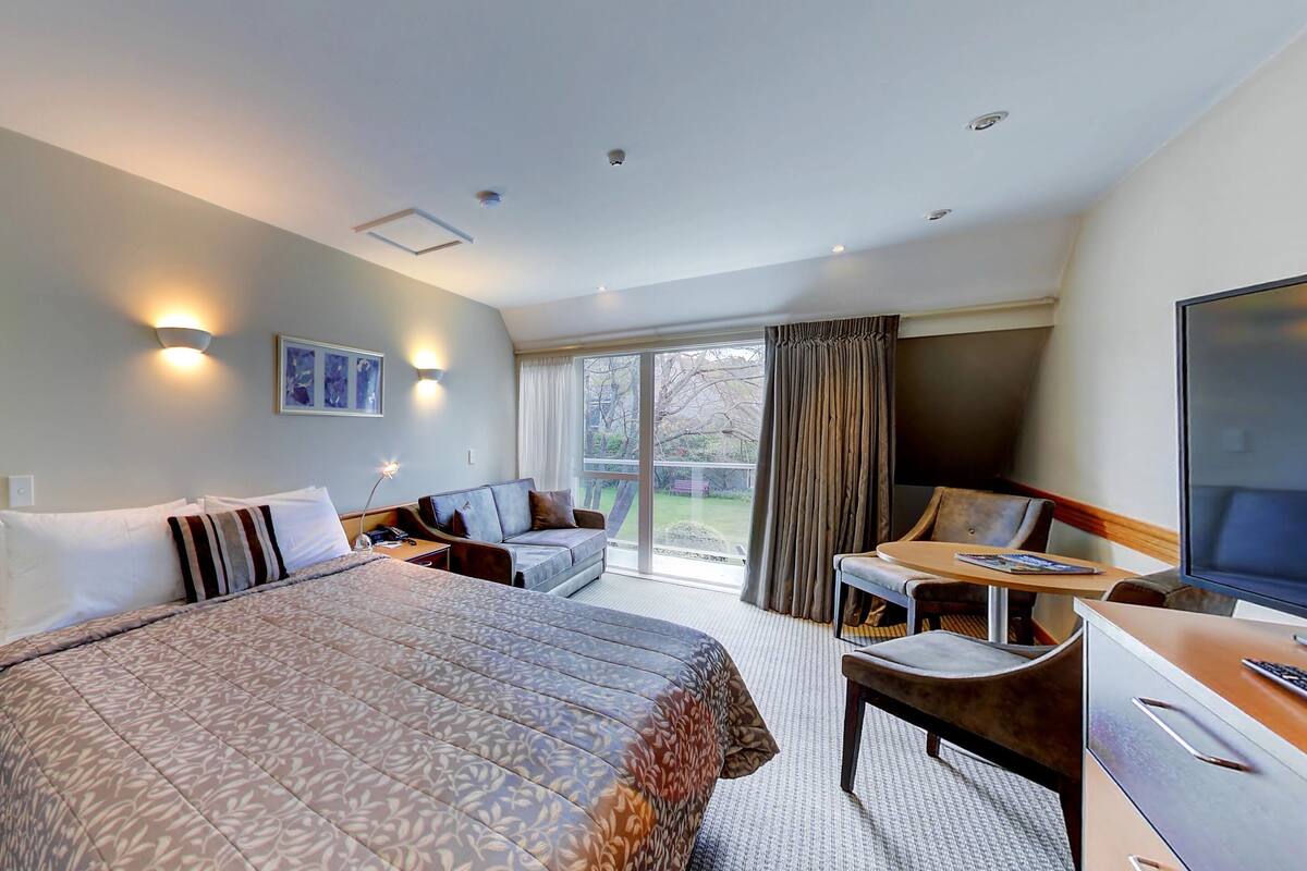 A hotel room with a large bed, a desk with a chair, a television, and a balcony with curtains.