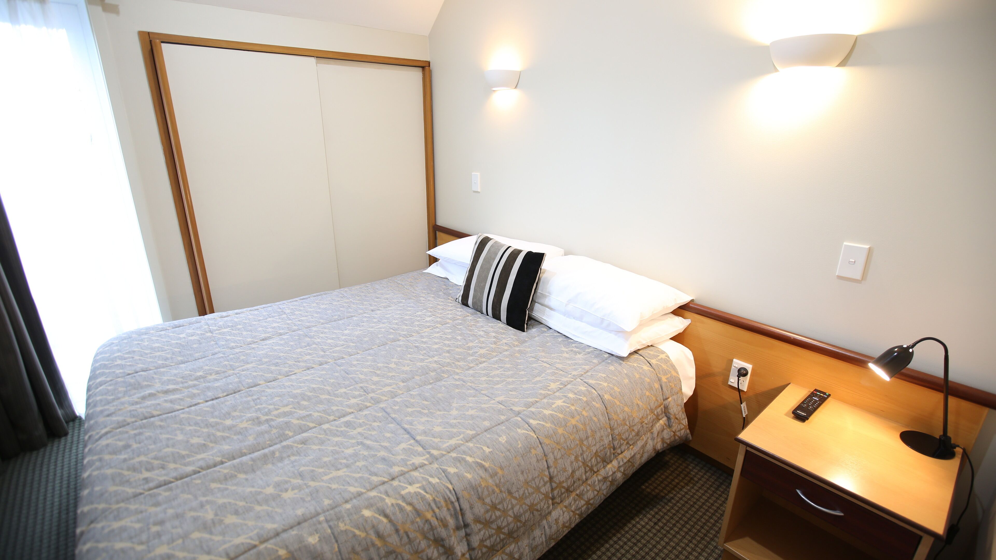 A hotel room with a bed, a nightstand, a lamp, and a wall-mounted light fixture.