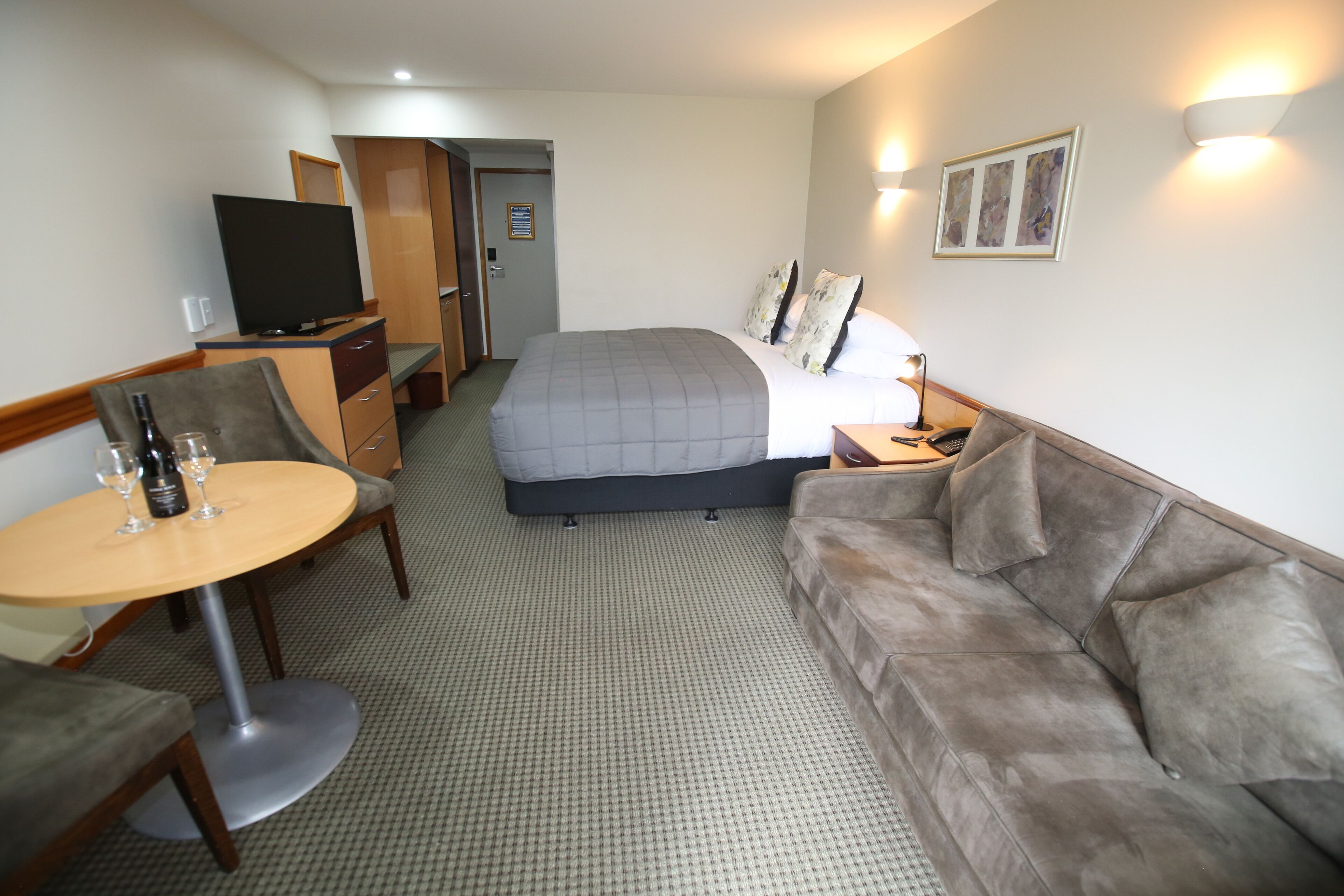 A hotel room with a bed, a sofa, a TV, a desk with a chair, and a small table with glasses and a bottle.