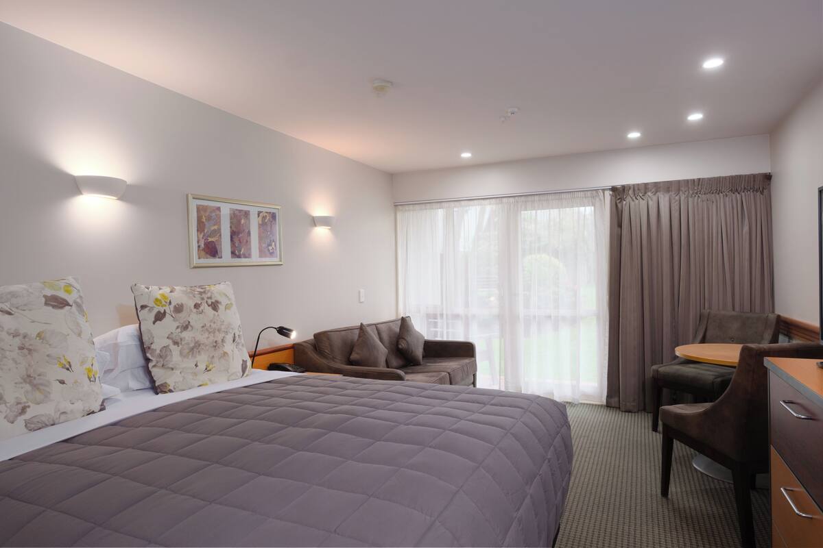 A hotel room with a large bed, a sofa, a desk, and a chair. There is a window with curtains and a painting on the wall.