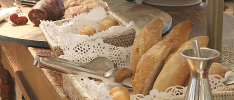 Variedad de panes y pasteles en una barra, incluyendo baguettes y croissants, con un colador metálico y un bol de frutas al fondo.
