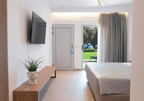 Un dormitorio moderno con una cama, un televisor montado en la pared, un banco con una planta y una puerta que da a un balcón con vistas a zonas verdes.