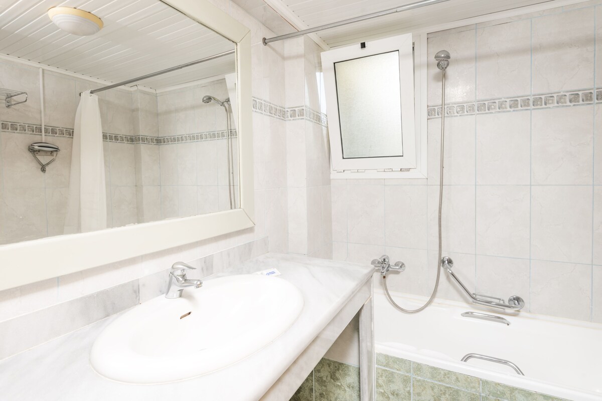 A bathroom with a white sink, a mirror, a shower with a handheld showerhead, and a tiled wall.