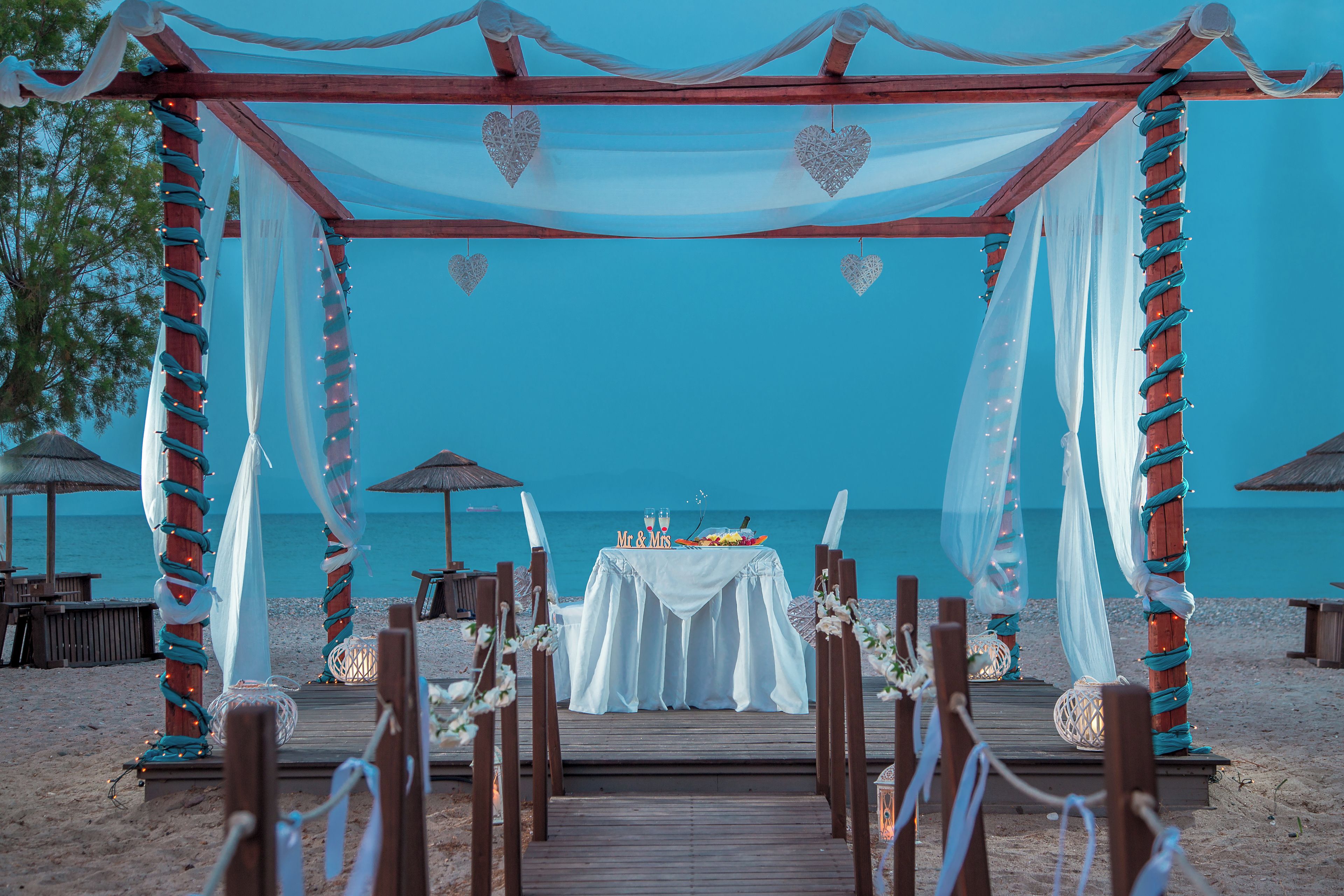Allestimento nuziale sulla spiaggia, con arco in legno, drappeggi bianchi e un tavolo con torta e bicchieri.