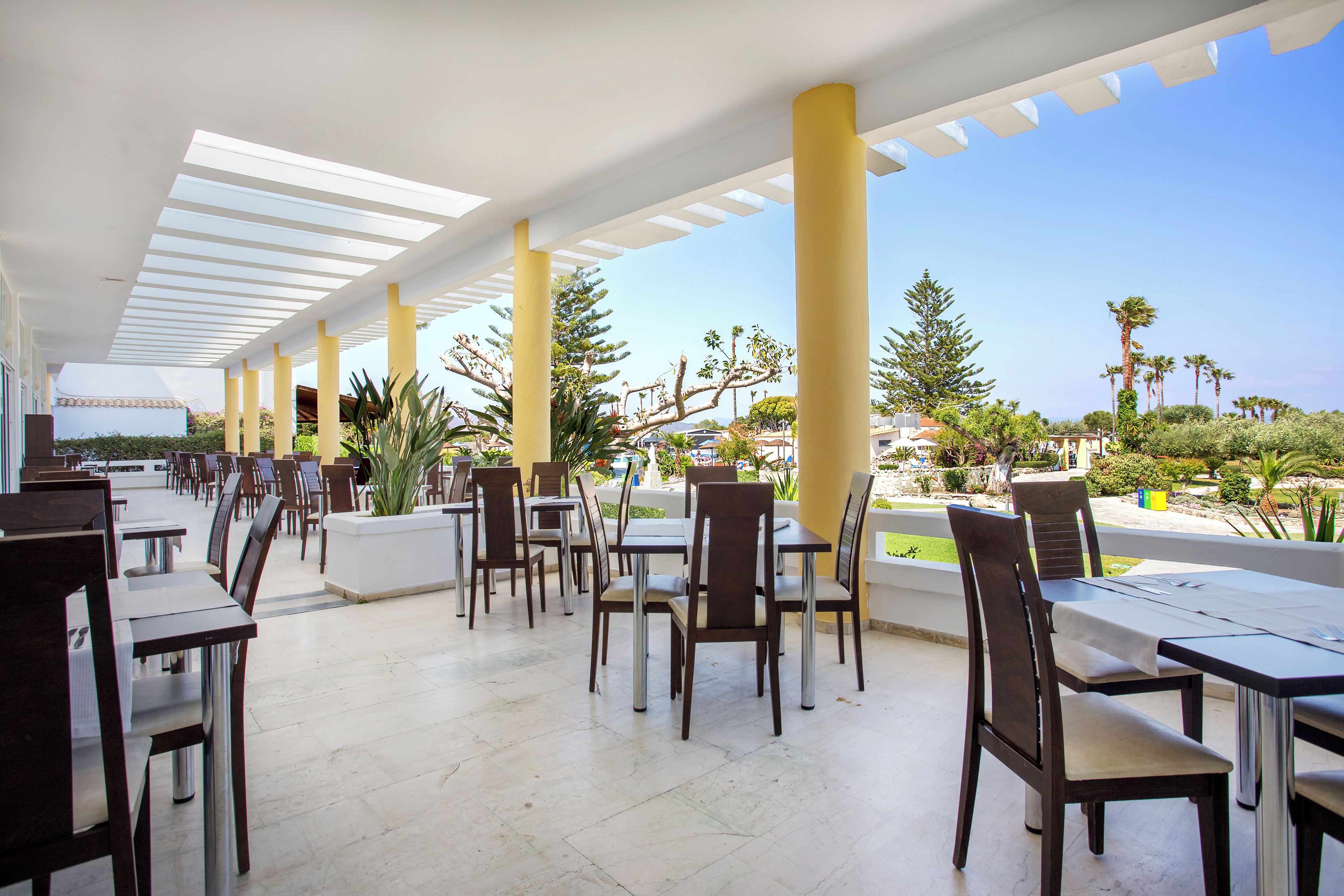 Un área de comedor al aire libre con varias mesas y sillas, un techo blanco con tragaluces y vistas a palmeras y follaje.