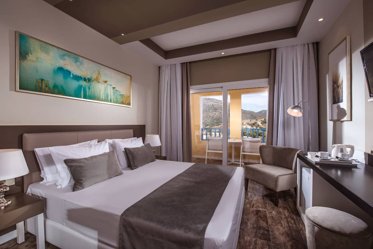 A hotel room with a large bed, a desk, a chair, and a balcony with a view of the mountains.