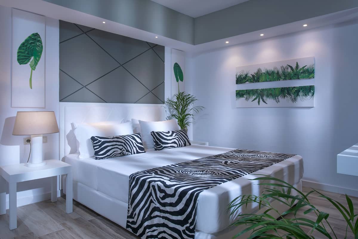 A modern bedroom with a large bed, white nightstands, a zebra-patterned bedspread, and wall art including a large leaf and a plant wall.