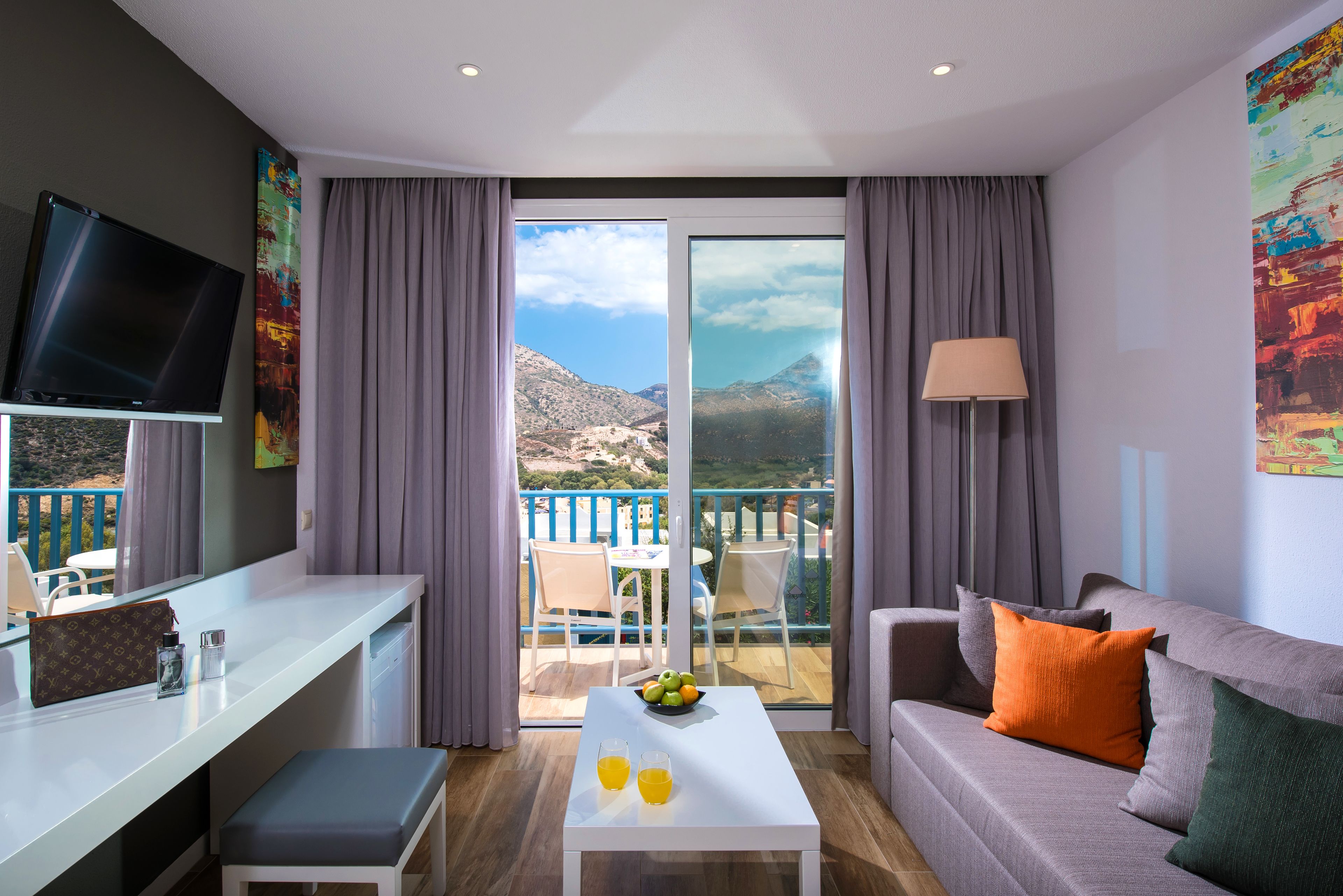 A modern hotel room with a balcony, a flat-screen TV, a dining table with fruit, and a sofa with cushions.