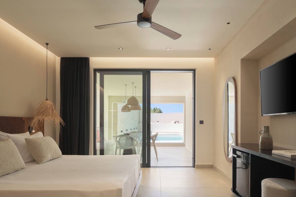 A modern hotel room with a bed, a ceiling fan, a TV, and a view of the beach through the door.