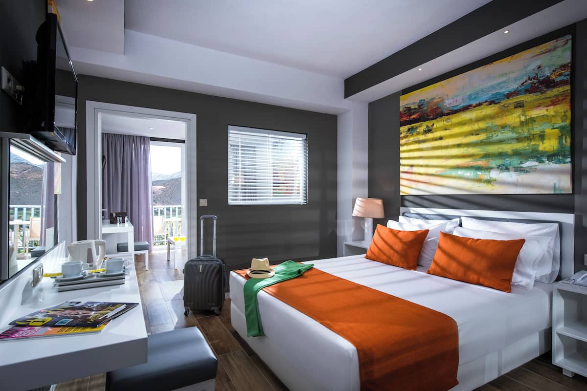 A hotel room with a bed, a desk, a chair, a suitcase, a window with curtains, and a painting on the wall.