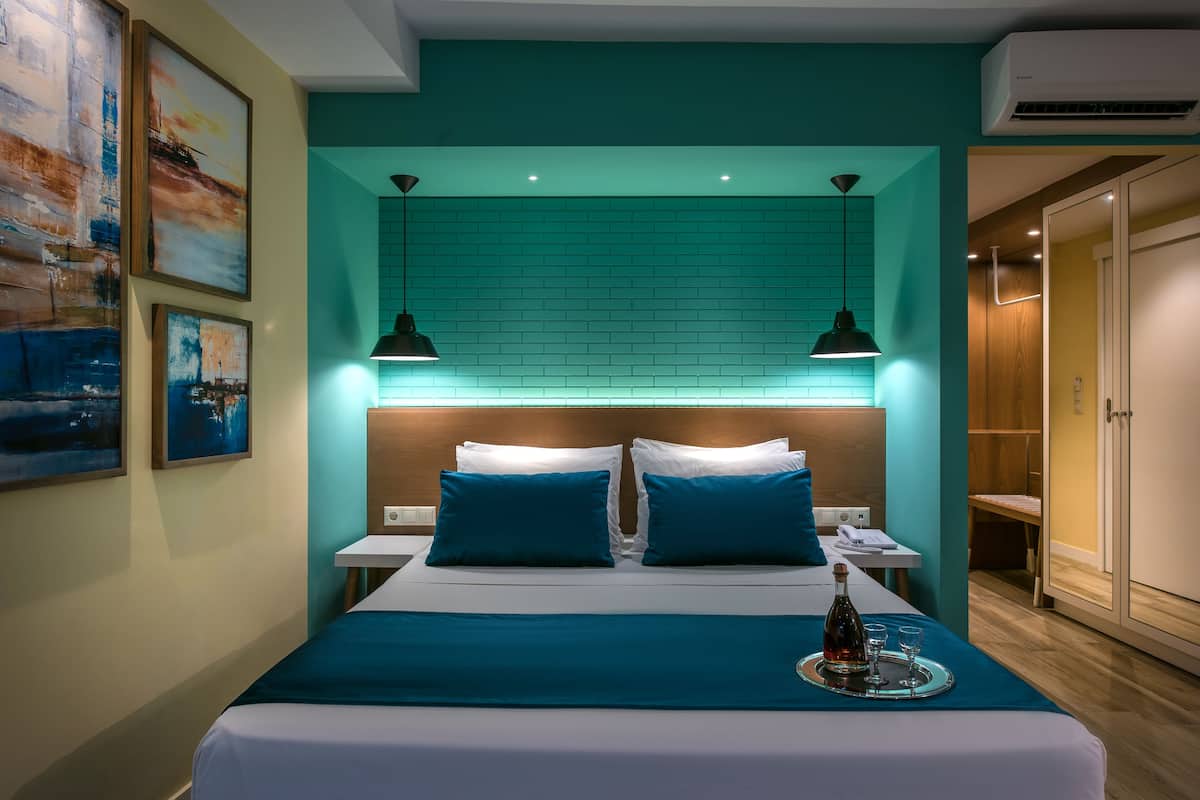 A modern hotel room with a teal accent wall, a large bed with blue bedding, two bedside lamps, and a tray with a bottle and glasses on the bed.