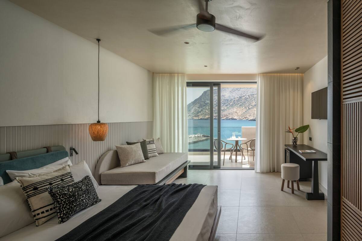 A modern hotel room with a bed, a desk, and a balcony with a view of the sea and mountains.
