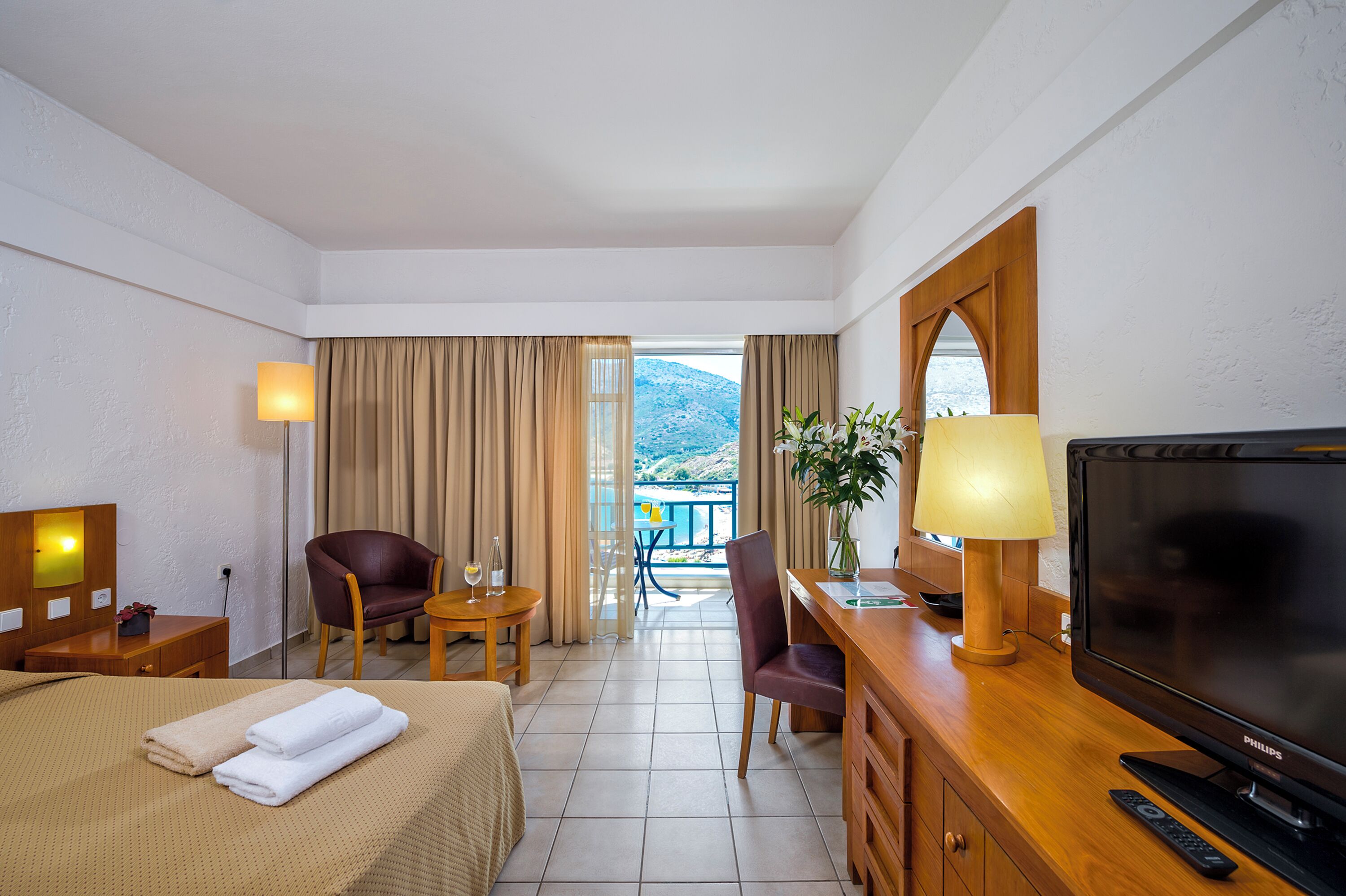 A hotel room with a bed, a desk, a TV, a chair, a lamp, and a view of the mountains.