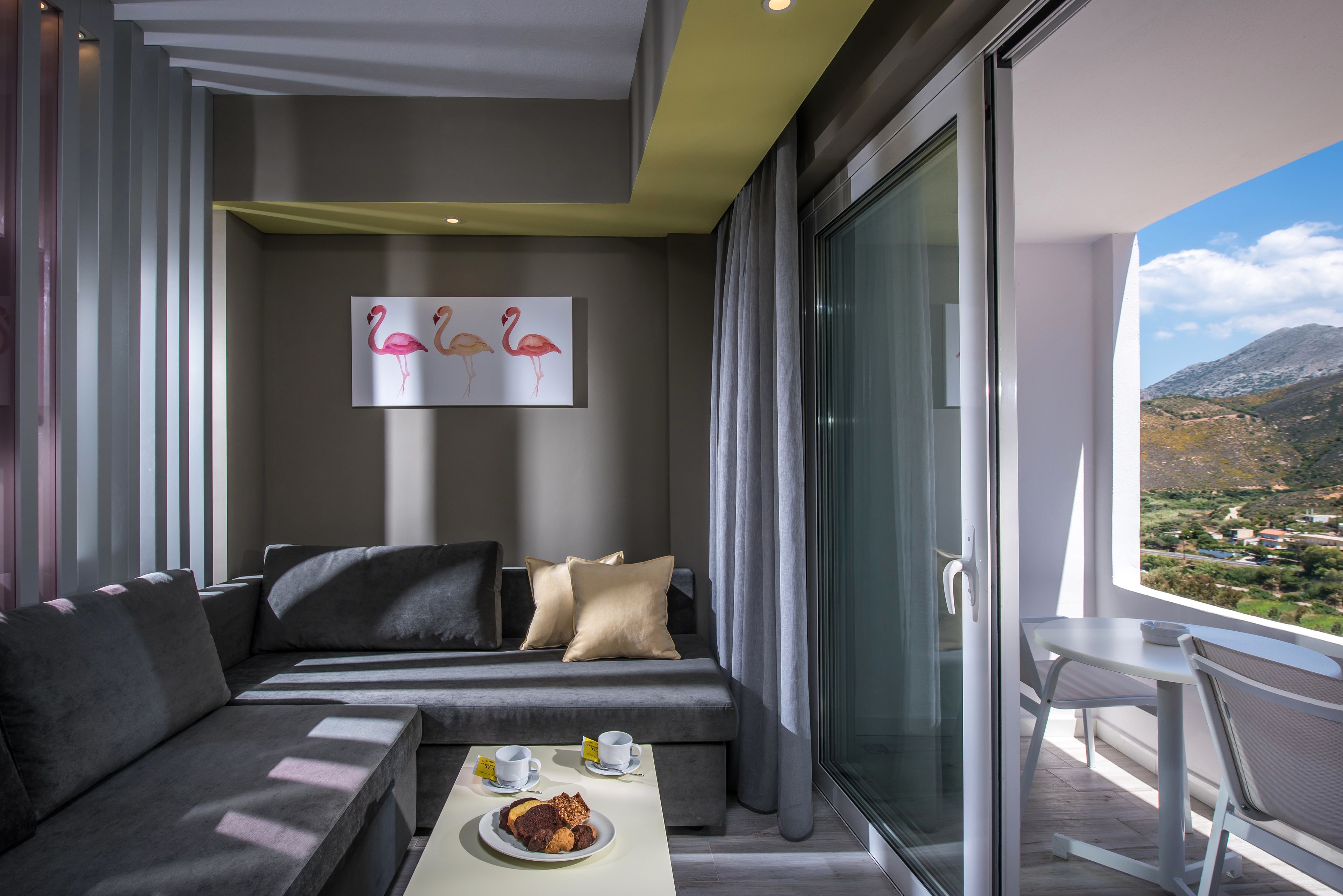 A modern hotel room with a sofa, a dining table with a plate of food, and a painting of flamingos on the wall.