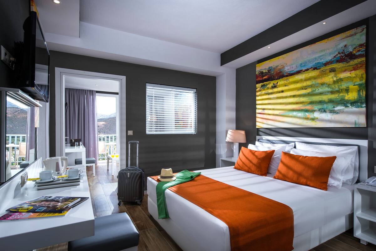 A hotel room with a bed, a desk, a chair, a suitcase, a window with curtains, and a painting on the wall.
